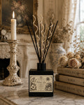 Island Lace Luxury Reed Diffuser matte black bottle with natural spiral and black reeds on French marble beside antique books and a taper candle